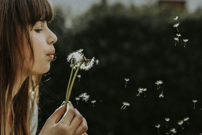 Inhouse Pharmacy’s Guide to Asthma Signs & Symptoms 1 A woman blowing a dandelion puff, sending its spores into the air.